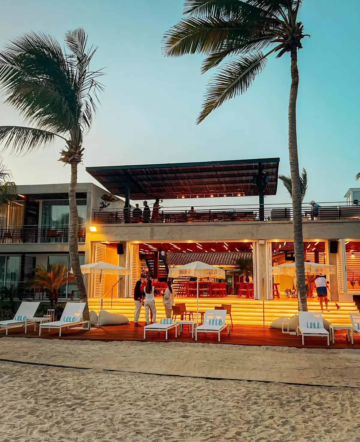 Edificio frente a la playa en el Gran Roque al anochecer, con la terraza y la planta baja iluminadas, palmeras y tumbonas en la arena